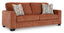 A rust-colored Ashley Aviemore Spice Sofa (2430438) featuring two seat cushions, two back cushions, and square armrests. This contemporary piece includes two decorative pillows with geometric patterns resting on the sofa.
