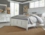 A bright bedroom with a large white bed, gray bedding, a jute rug, white dresser, lamps, a mirror, and sunlight streaming through tall windows with white curtains.