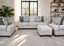 A living room featuring the Annisa Julien Cork Sofa and Loveseat by Franklin Furniture Corp. (957-3574-20) in light gray, designed with reversible cushions and gel-blended foam seating, complemented by floral and blue pillows, a matching ottoman, a bookshelf, potted plant, large windows with sheer curtains, and a light rug.