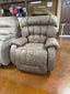 The Rake Mushroom Recliner 9B14-22389 by Best Home Furnishings—a plush, oversized recliner with thick cushioning—sits on a shiny brown floor in a furniture store, positioned near another similar chair and shelves stocked with fabric rolls in the background.