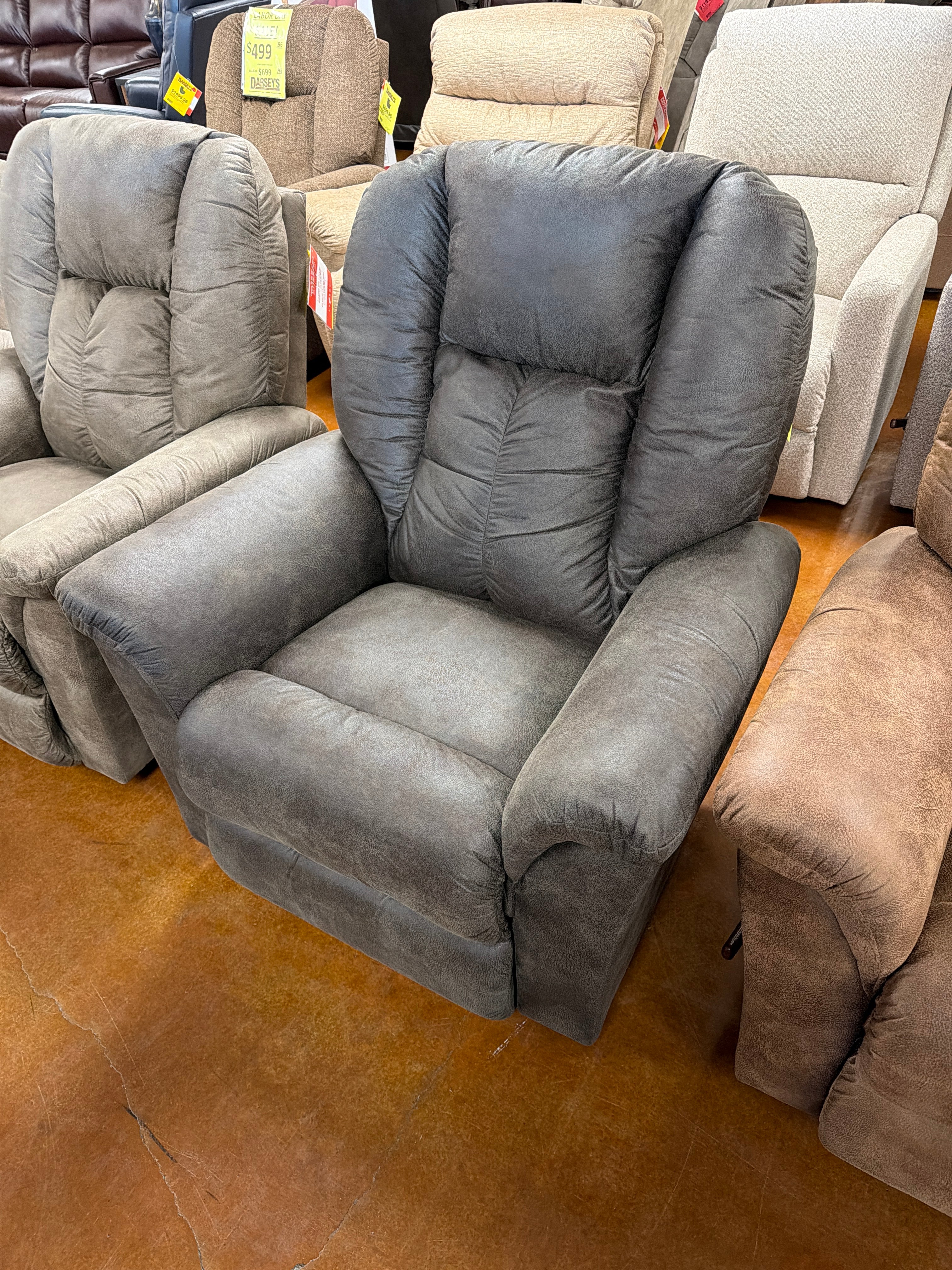 A La-Z-Boy Jasper Recliner 709-10-D1825-58 in plush gray is showcased on a brown floor among other recliners in a furniture store. With its ultra-plush chaise seat, padded arms and backrest, and wide recliner design, this chair delivers exceptional comfort for relaxing at home.