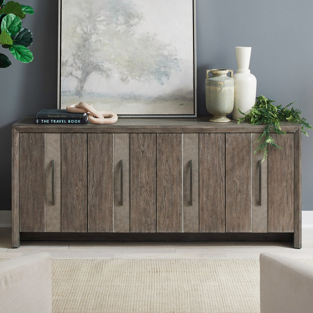 The Lafayette Curio Cabinet 2219-AC1000 by Liberty Furniture, featuring elegant vertical handles, is styled as an accent piece and topped with decorative vases, a plant, books, and a framed landscape painting. Positioned against a gray wall and set atop a cream rug, this cabinet serves as both a stylish storage solution and a striking focal point in the room.