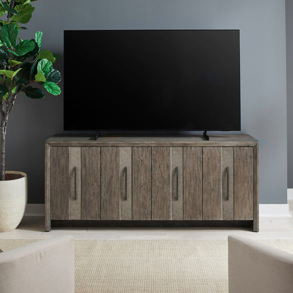 The Lafayette Curio Cabinet 2219-AC1000 by Liberty Furniture stands beneath a flat-screen TV, its elegant wooden design with vertical handles providing an attractive storage solution. A potted plant is placed to the left, complementing the room’s neutral walls and light flooring.