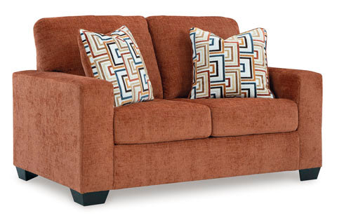 The Ashley Aviemore Spice Loveseat 2430435 is a contemporary loveseat in a rich rust shade, featuring wide arms and two patterned throw pillows with geometric designs in brown, blue, and cream.