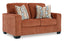 The Ashley Aviemore Spice Loveseat 2430435 is a contemporary loveseat in a rich rust shade, featuring wide arms and two patterned throw pillows with geometric designs in brown, blue, and cream.