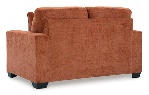 The Ashley Aviemore Spice Loveseat 2430435 is shown from the back, featuring a burnt orange upholstered contemporary design with square arms, plush cushions, and short black feet against a white background.