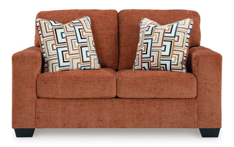 The Ashley Aviemore Spice Loveseat 2430435 is a contemporary burnt orange loveseat with wide arms and dark wooden legs, featuring two square patterned pillows with geometric designs in beige, black, blue, and brown.