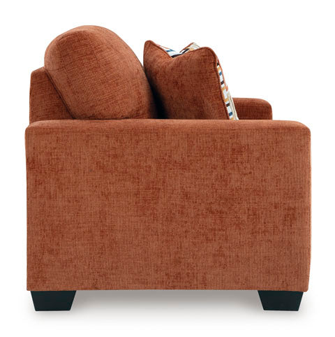 Side view of the Ashley Aviemore Spice Loveseat 2430435, a modern burnt orange loveseat featuring textured fabric, square arms, black feet, and two cushions—one solid and one with a patterned edge visible behind the arm.
