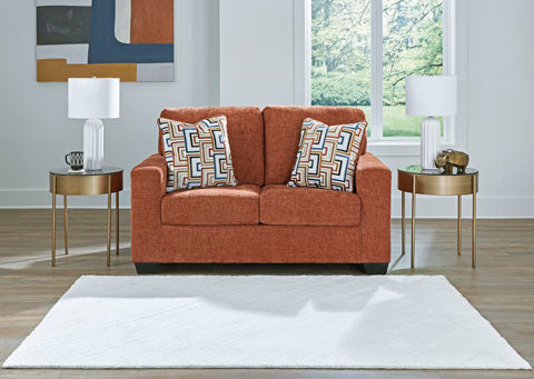 The Ashley Aviemore Spice Loveseat 2430435, upholstered in a vibrant burnt orange shade, is paired with two geometric-patterned pillows and flanked by round side tables crowned with lamps. Situated in a bright room adorned with a window, abstract artwork, and a white rug atop wooden flooring, this stylish setup radiates modern appeal.