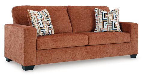 A rust-colored Ashley Aviemore Spice Sofa (2430438) featuring two seat cushions, two back cushions, and square armrests. This contemporary piece includes two decorative pillows with geometric patterns resting on the sofa.