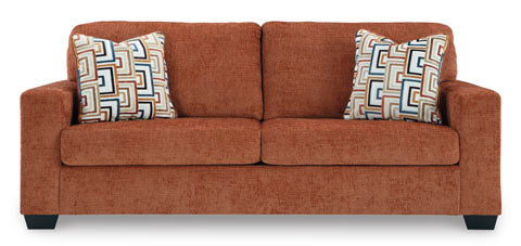 The Ashley Aviemore Spice Sofa 2430438 is a contemporary sofa upholstered in rust-colored fabric, featuring square arms and two geometric-patterned throw pillows, displayed against a plain white background.