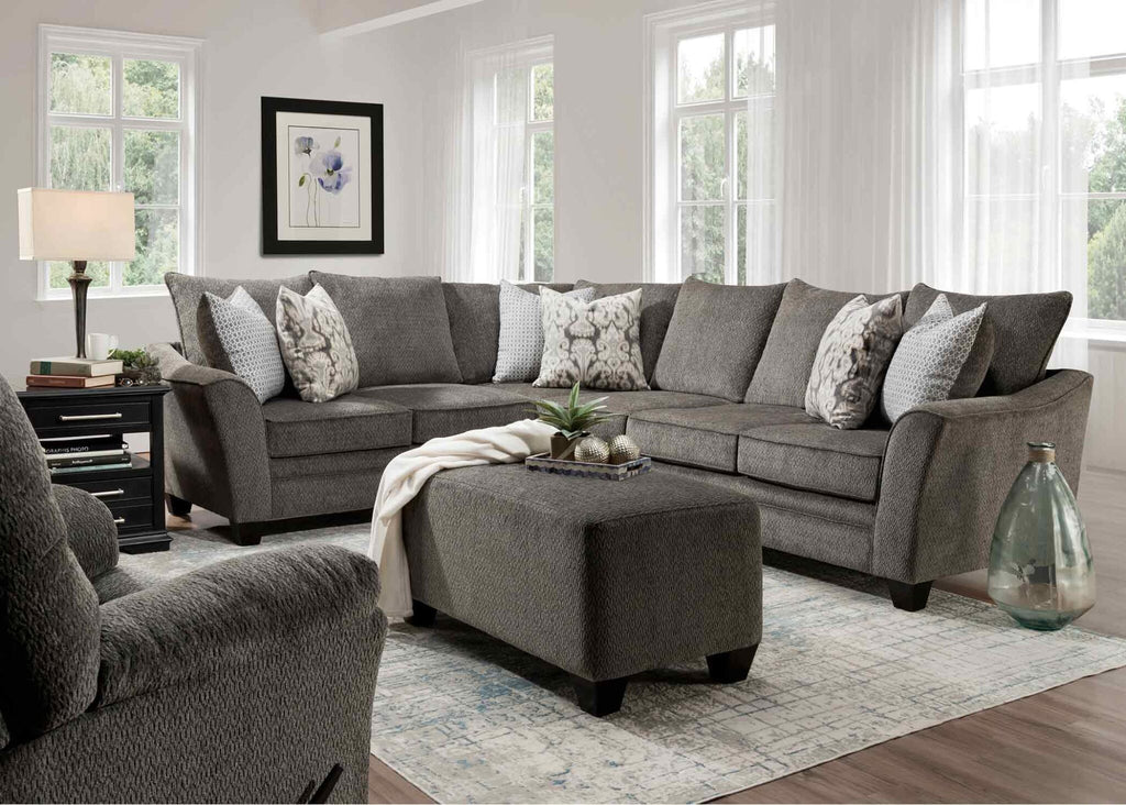 A modern living room with a spacious Fairmont Shasta Charcoal Sectional by Franklin Furniture Corp. (256-28-49-3403-05), accented with patterned pillows, an upholstered ottoman, a lamp, a framed flower print, and a glass vase on a rug near bright windows draped with sheer curtains.