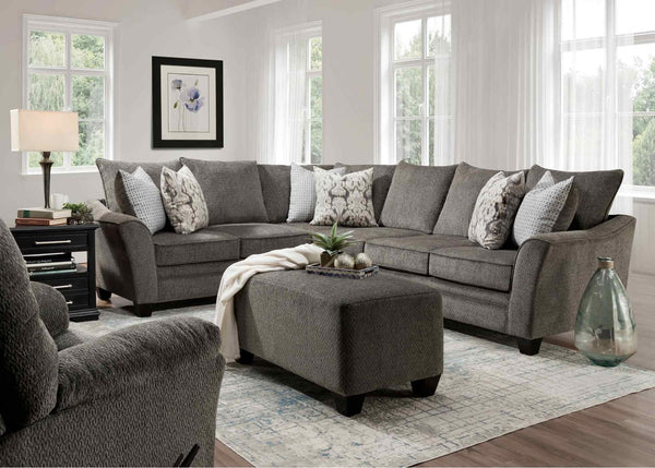 A modern living room with a spacious Fairmont Shasta Charcoal Sectional by Franklin Furniture Corp. (256-28-49-3403-05), accented with patterned pillows, an upholstered ottoman, a lamp, a framed flower print, and a glass vase on a rug near bright windows draped with sheer curtains.