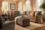 A cozy living room featuring the Fairmont Shasta Charcoal Sectional by Franklin Furniture Corp., patterned pillows, an oversized cocktail ottoman, a beige rug, a potted plant, framed art, and sunlight streaming through multiple windows with sheer curtains.