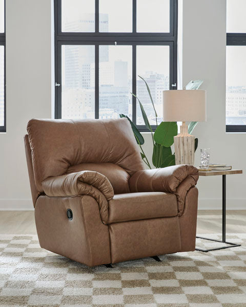 The Ashley WillowBend Caramel Recliner 2770425 is placed on a patterned rug beside a side table with a lamp and glass, in a bright room featuring large windows and city views.
