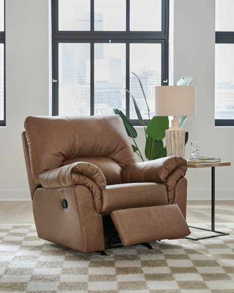 A WillowBend Caramel Recliner 2770425 by Ashley, upholstered in brown leather and featuring an extended footrest, sits on a checkered rug. Behind it is a table with a lamp, a plant, and large windows displaying a cityscape.