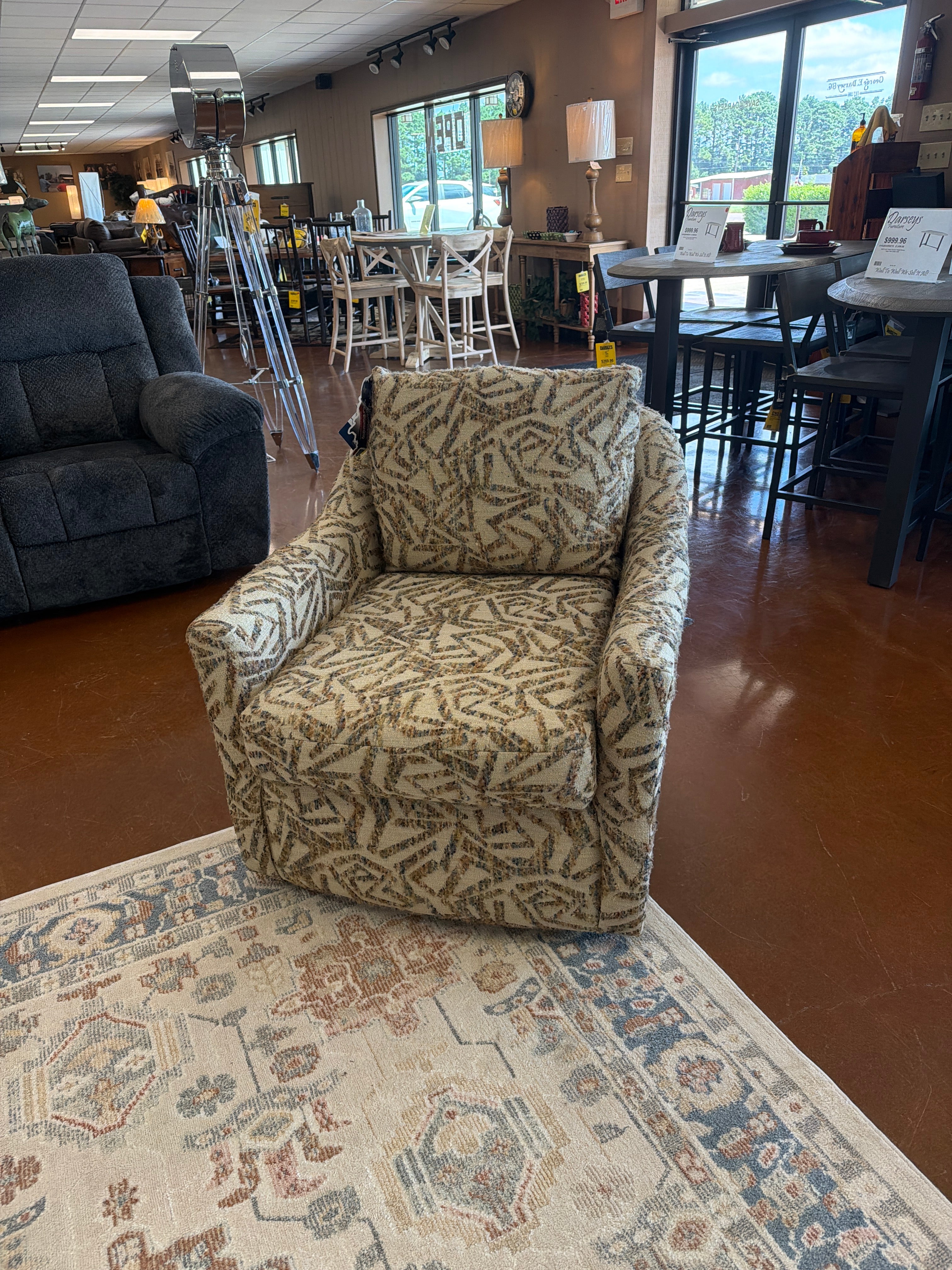 The Praslin Multi Swivel Chair 2328-35919 by Best Home Furnishings, featuring earth-toned geometric upholstery and a plush pillow back, is displayed on a cream and blue rug in the furniture store, surrounded by other chairs, tables, and large windows.