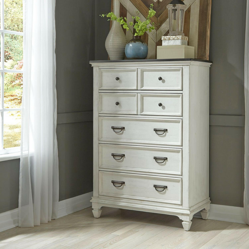 A white wooden dresser with five drawers stands in a corner near a window with sheer curtains. Decorative items, including vases and a lantern, are arranged on top.