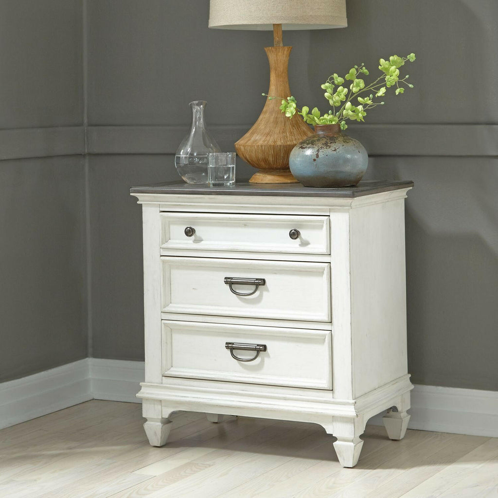 A white wooden nightstand with three drawers sits in a corner, topped with a lamp, a glass decanter, and a ceramic vase containing green flowers. The walls are gray and the floor is light wood.