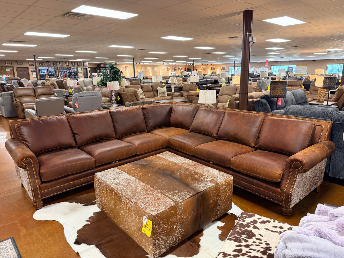 Preserve Caramel Longhorn Dark Sectional 4300LH70-14-15 L-shaped brown leather sectional with nailhead trim and a square cowhide-patterned ottoman on a cowhide rug in a furniture showroom.