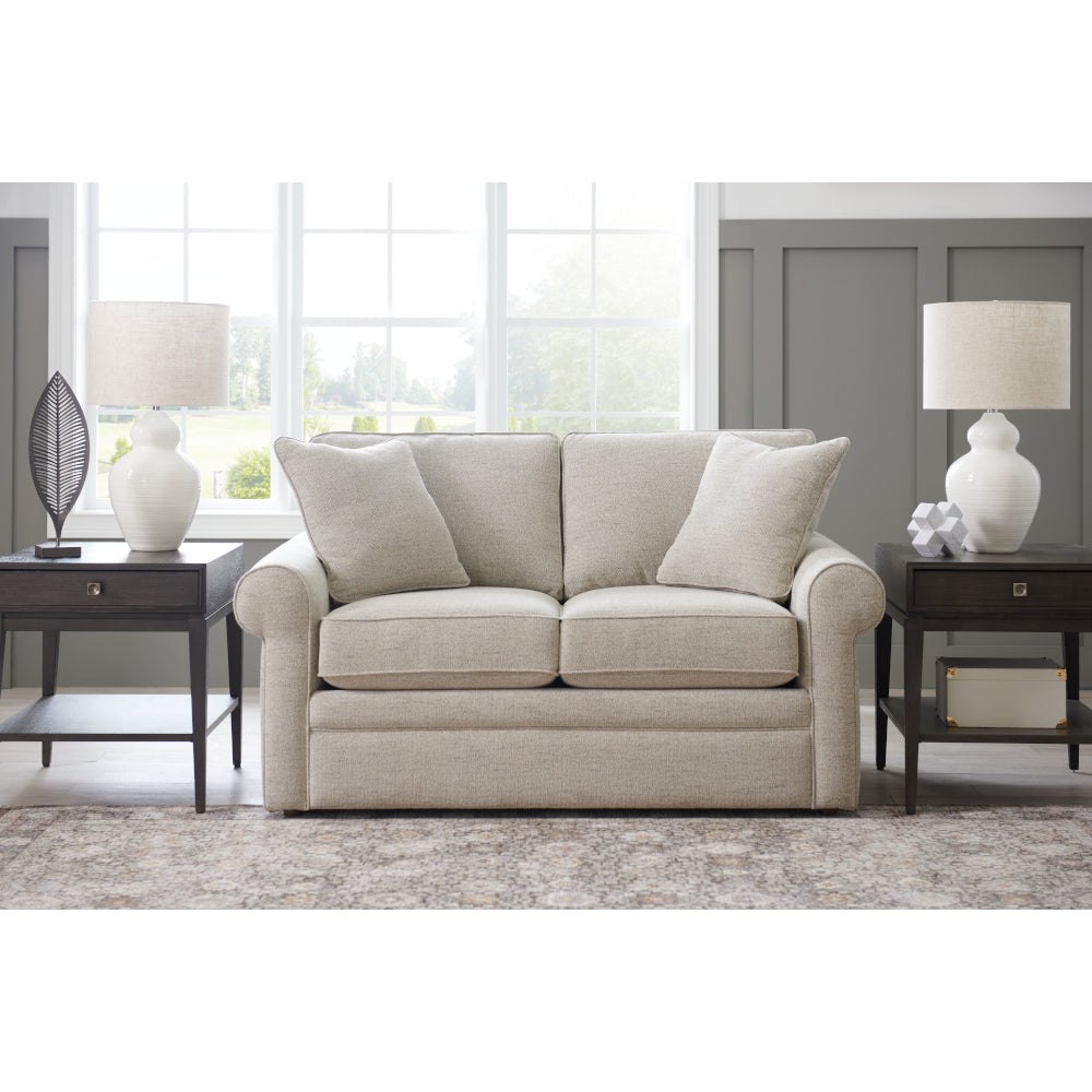 A La-Z-Boy Collins Loveseat 494-630-D1807-64, featuring box seat cushions and beige upholstery, is centered between two dark wood side tables topped with white lamps in a bright living room with large windows and gray paneled walls.