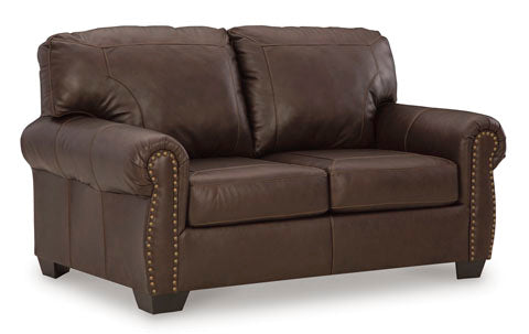 The Ashley Colleton Loveseat 5210735 showcases dark brown leather upholstery, rolled arms with nailhead trim, and two seat cushions, all set against a plain white background for a timeless traditional appeal.
