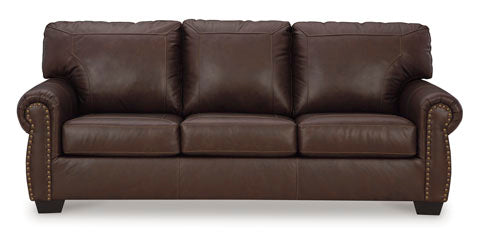 The Ashley Colleton Sofa 5210738 features dark brown leather upholstery, three seat cushions, three back cushions, rolled arms enhanced with brass nailhead trim, and short wooden legs. Shown against a white background, this classic traditional sofa adds timeless elegance to any living space.