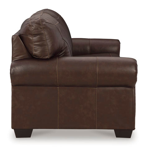 Side view of the Ashley Colleton Sofa 5210738 in dark brown leather, featuring visible stitching, wide roll armrests in a classic sofa style, a cushioned back, and short wooden legs, set against a white background.