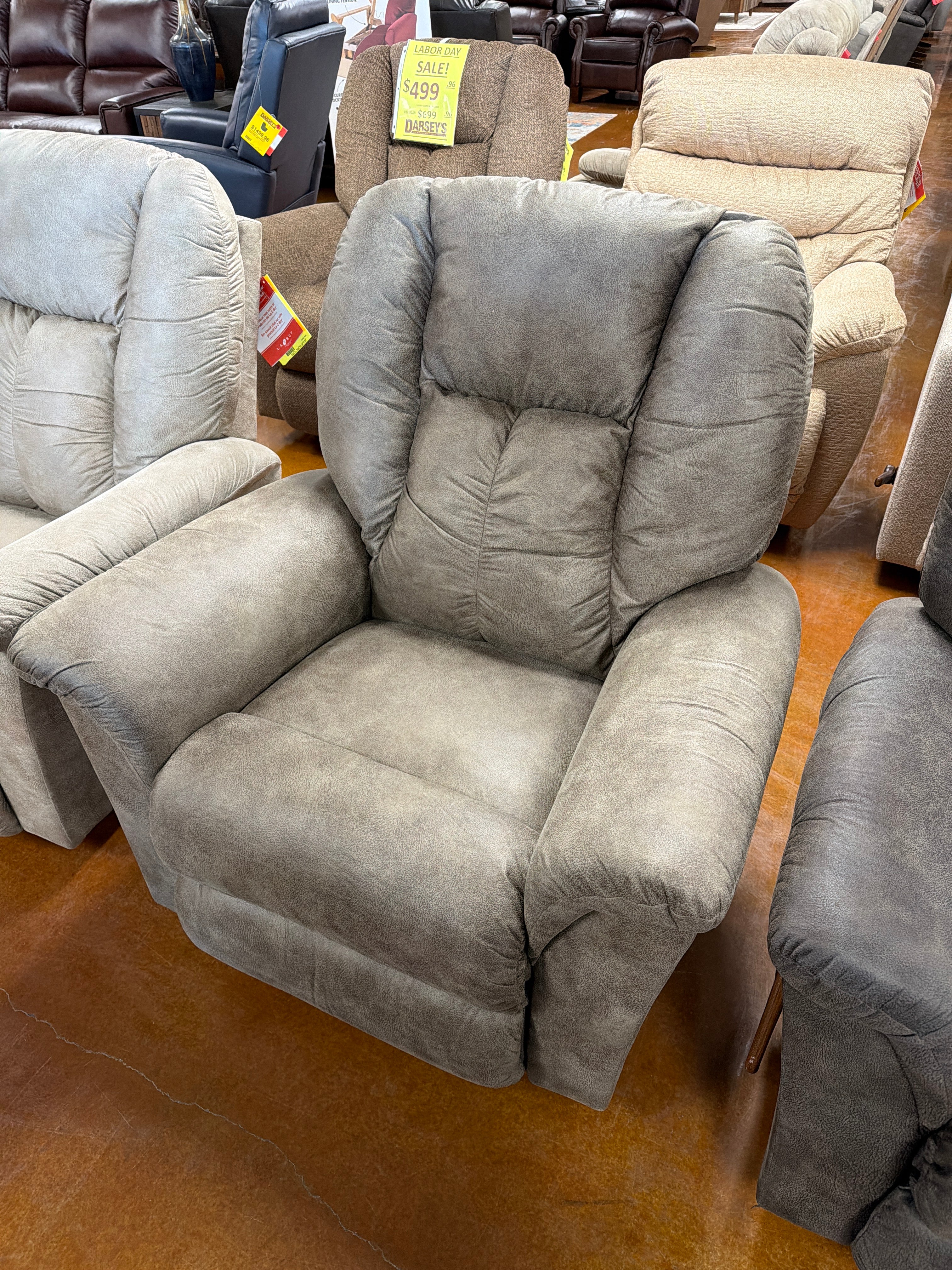 A large, gray upholstered Jasper Recliner 709-10-D1825-73 by La-Z-Boy is displayed in a furniture showroom alongside similar chairs. A yellow sale tag is visible on another chair in the background.