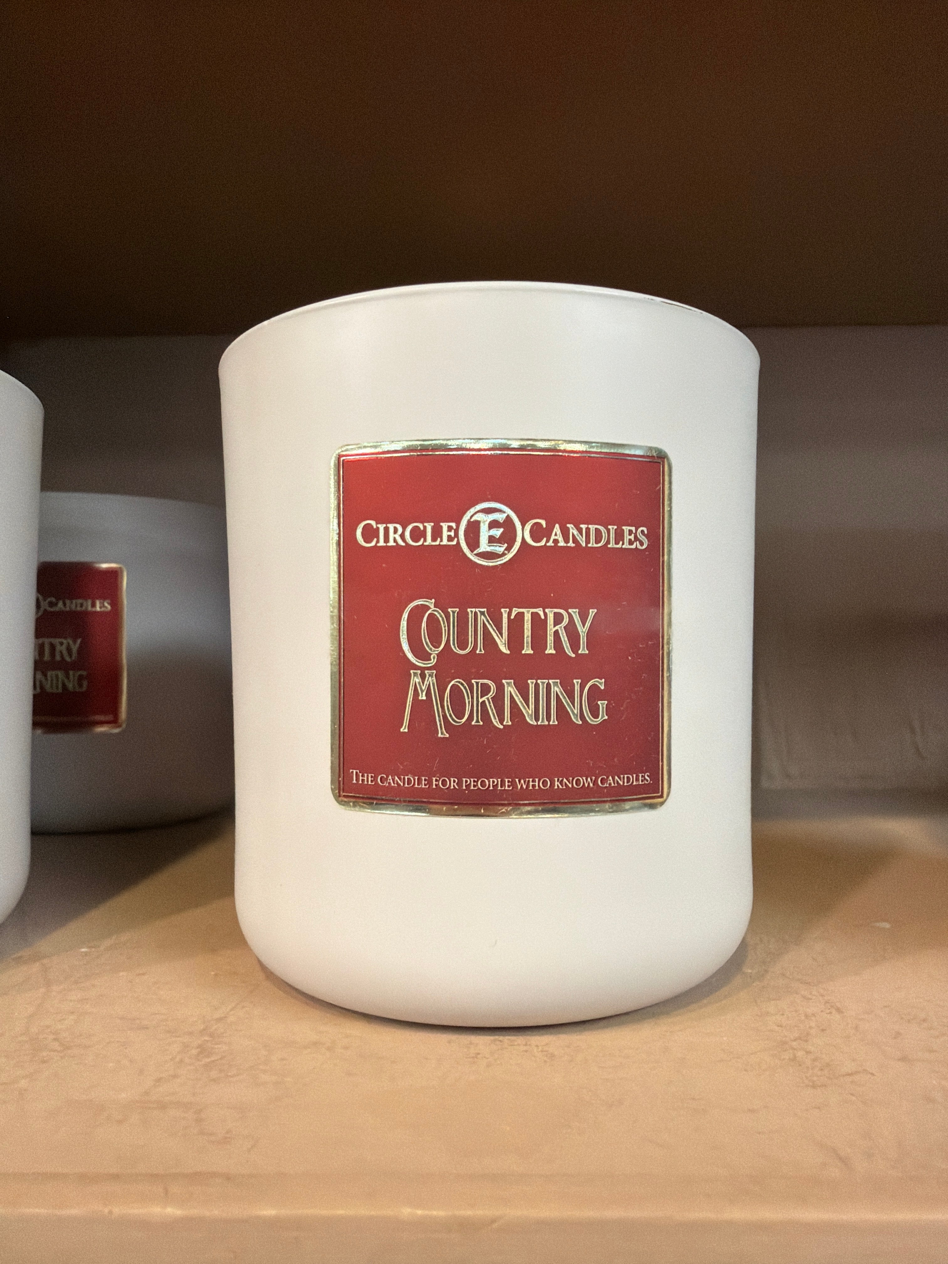 A white jar labeled Circle E Candle 14oz Country Morning rests on a brown shelf, filling the air with subtle notes of vanilla and nutmeg. The label reads, “The candle for people who know candles.” Another identical jar is partially visible in the background.