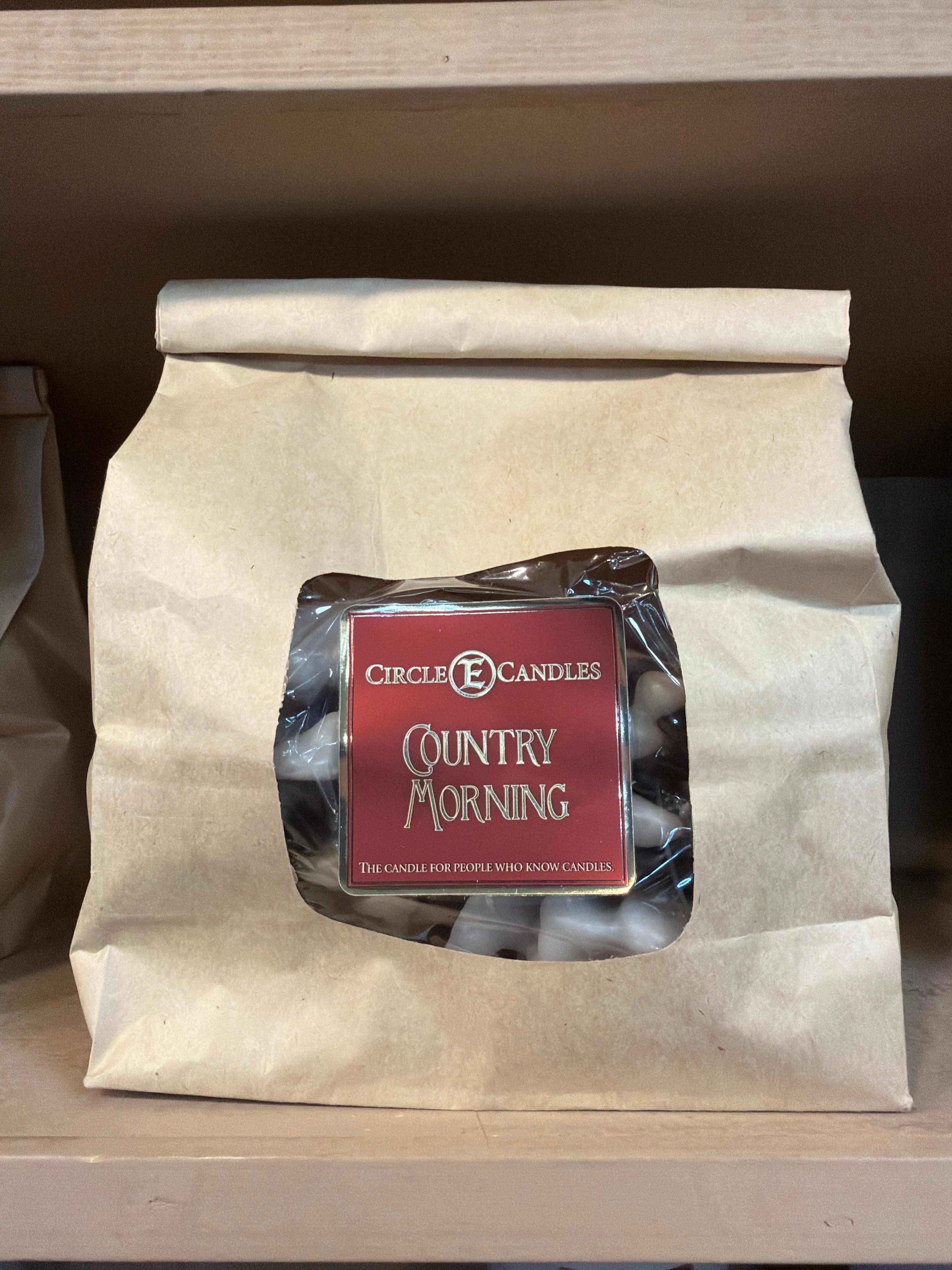 A brown paper bag with a red label that reads Circle E Candle Firestarters Country Morning, featuring warm notes of cinnamon pumpkin spice, sitting on a shelf.