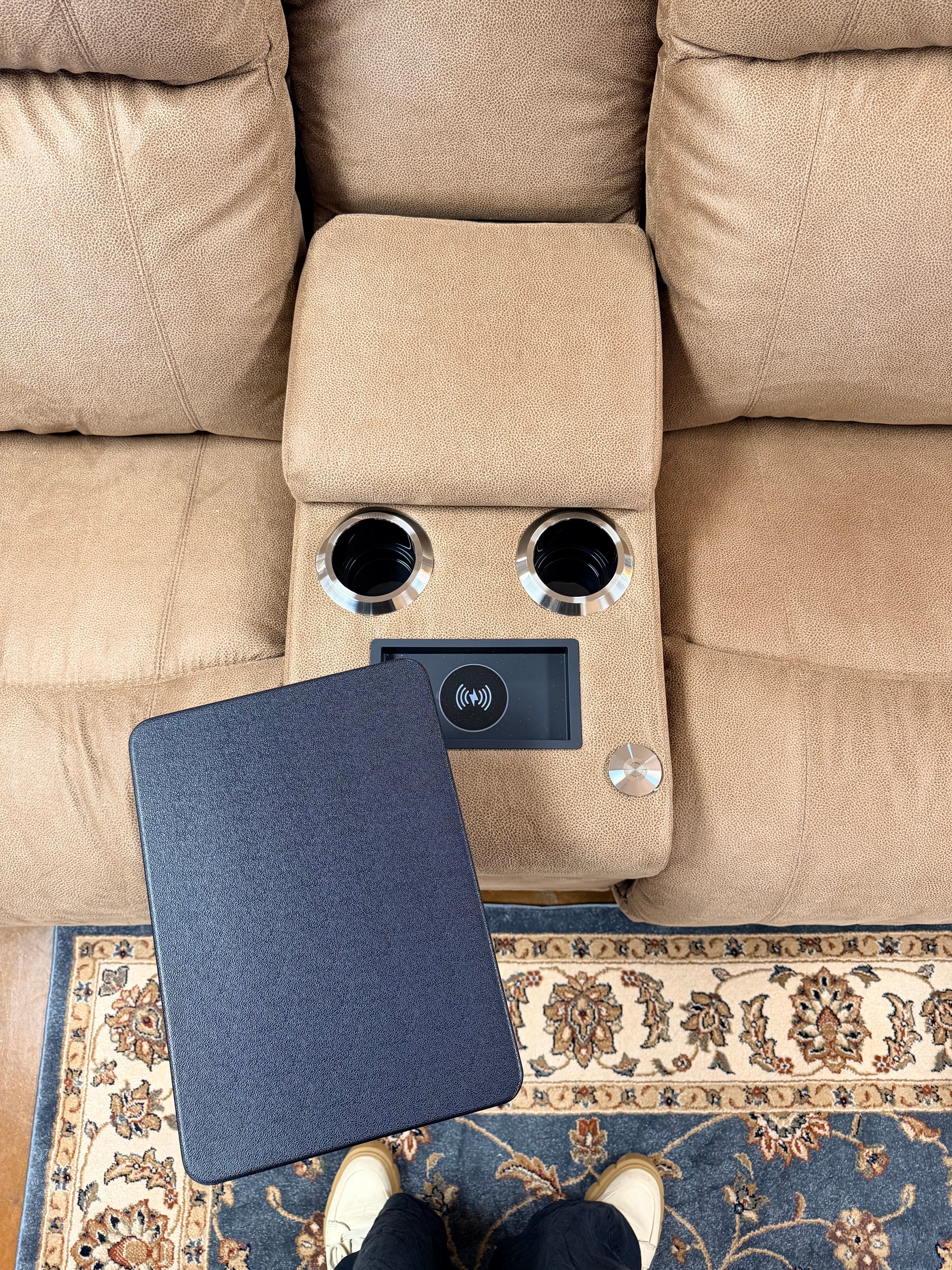 An overhead view of the Dorian Power Loveseat Reclining 755-X49-D1855-75 in tan, featuring a built-in console with two cup holders, a wireless charging pad, and removable black tray table. A patterned rug and part of a person’s shoes appear at the bottom.