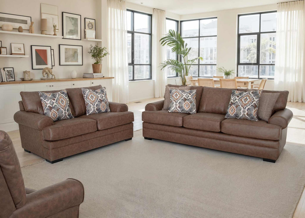 A modern living room features the Franklin Furniture Corp. Langston Duvall Vintage Brown Sofa and Loveseat 832-3429-16 upholstered in fabric, complemented by a beige rug and patterned throw pillows. Large windows bathe the space in natural light, while a dining table, lush plants, and wall art enhance the bright, airy ambiance. Gel-blended foam seating ensures exceptional comfort throughout the room.