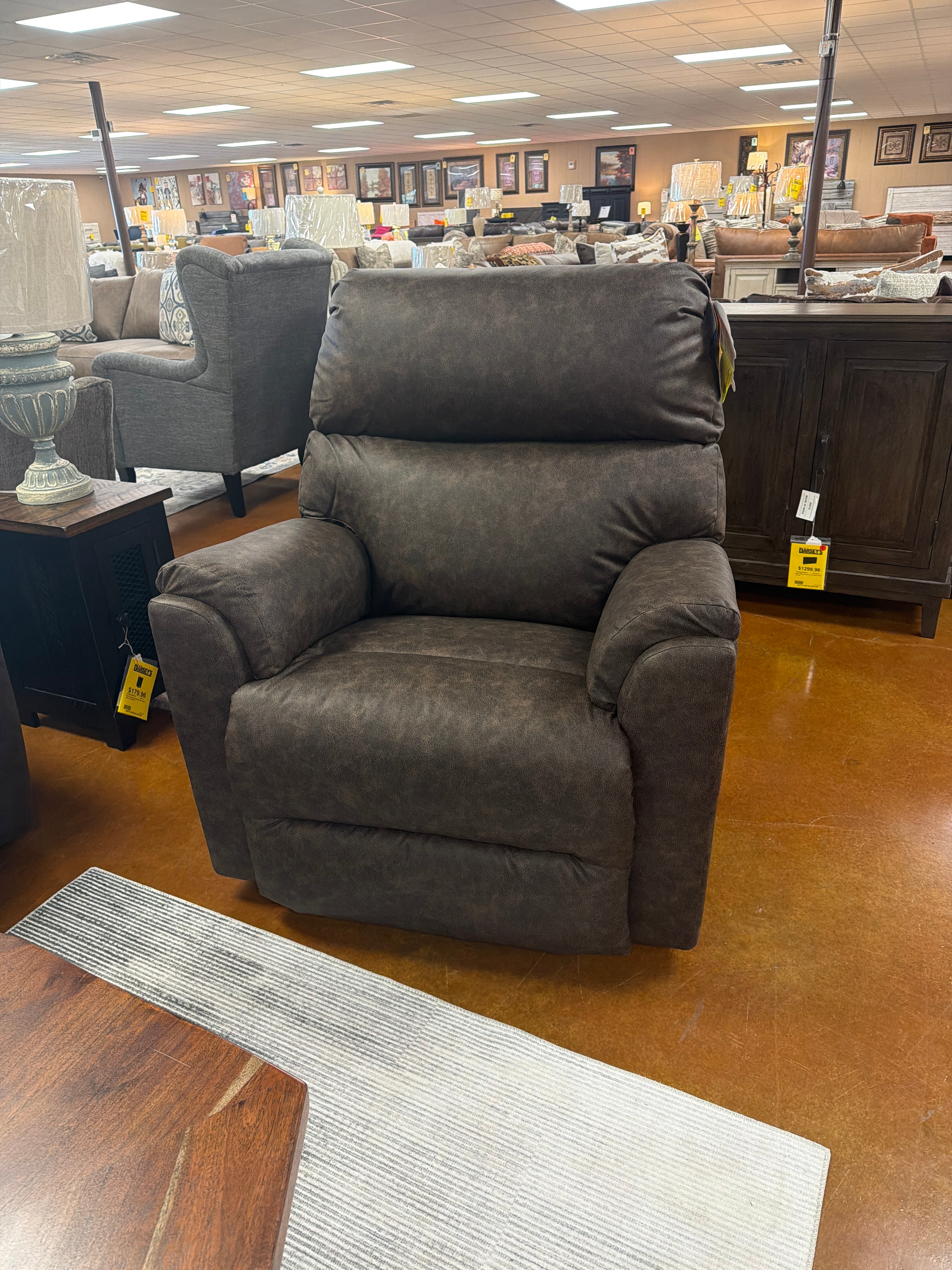 The Conquistador Walnut Recliner 9B97-22386 by Best Home Furnishings—a heavy-duty dark brown upholstered chair—sits on a light rug in a furniture showroom, surrounded by sofas, lamps, and wooden furniture under bright ceiling lights.