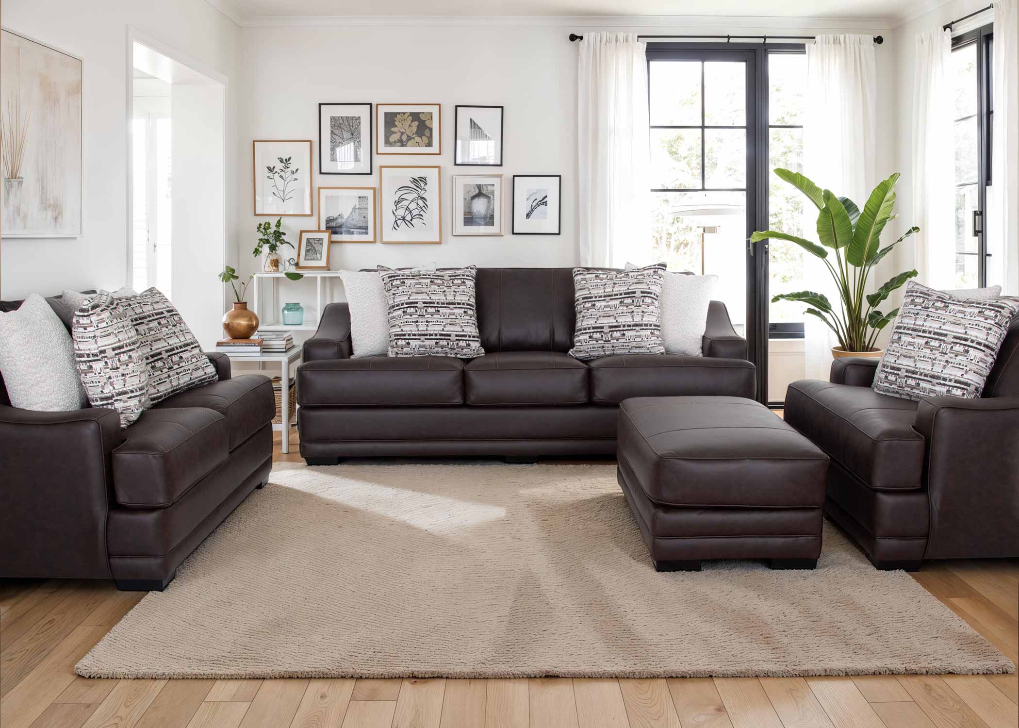A modern living room showcases the Prescott Duvall Coffee Sofa and Loveseat 893-3429-10 by Franklin Furniture Corp., upholstered in rich dark brown leather. The seating is enhanced with patterned pillows and paired with a cozy beige rug, artistic wall decor, vibrant lush plants, and expansive windows dressed in white curtains that bathe the space in natural light. The included WOW Seating System elevates both comfort and style for a superior lounging experience.
