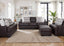 A modern living room showcases the Prescott Duvall Coffee Sofa and Loveseat 893-3429-10 by Franklin Furniture Corp., upholstered in rich dark brown leather. The seating is enhanced with patterned pillows and paired with a cozy beige rug, artistic wall decor, vibrant lush plants, and expansive windows dressed in white curtains that bathe the space in natural light. The included WOW Seating System elevates both comfort and style for a superior lounging experience.
