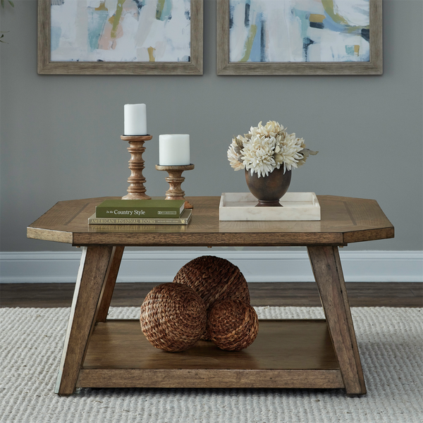 Here’s the rewritten sentence using your product data: The Liberty Furniture Carolina Park Cocktail Table 930-OT1010 features two candlesticks, books, and a flower arrangement arranged neatly on top, while woven decorative balls accent the lower shelf. Abstract artwork is displayed on the wall behind the table.