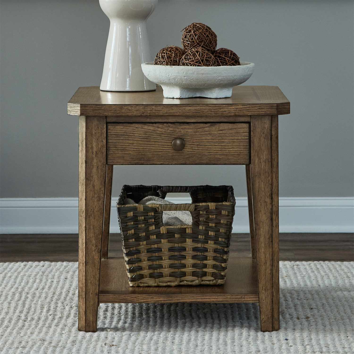 The Liberty Furniture Carolina Park End Table 930-OT1020, complete with a storage drawer and a woven basket on the lower shelf, is elegantly displayed on a textured light rug against a gray wall, adorned with a white vase and a decorative bowl filled with rattan balls.