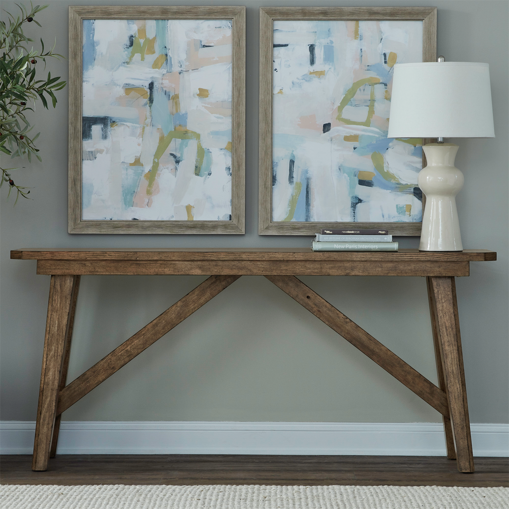 A Liberty Furniture Carolina Park Sofa Table 930-OT1030, crafted with white oak veneers and finished in rich Amber Brown, sits against a gray wall adorned with books and a white lamp. Above it hang two abstract paintings in soft pastel colors, while a plant’s leaves are partially visible on the left edge.