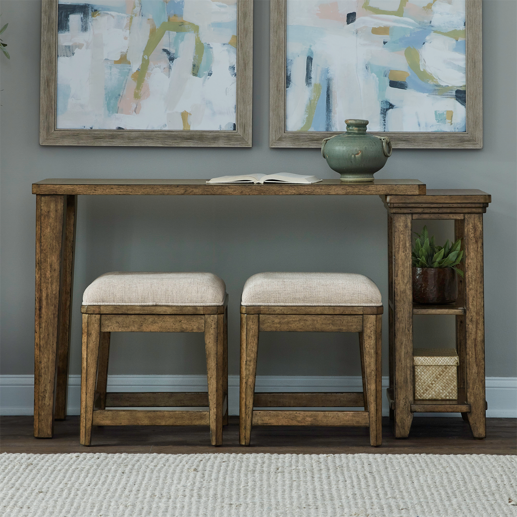 The Carolina Park Console Table and 2 Stools by Liberty Furniture offers elegant bookshelf storage, paired with two beige upholstered stools. Atop the wooden console table sits a green vase and an open book, while two abstract paintings above infuse artistic charm. A small side shelf holds a basket and another decorative pot, completing the look on a light rug for a cozy finish.