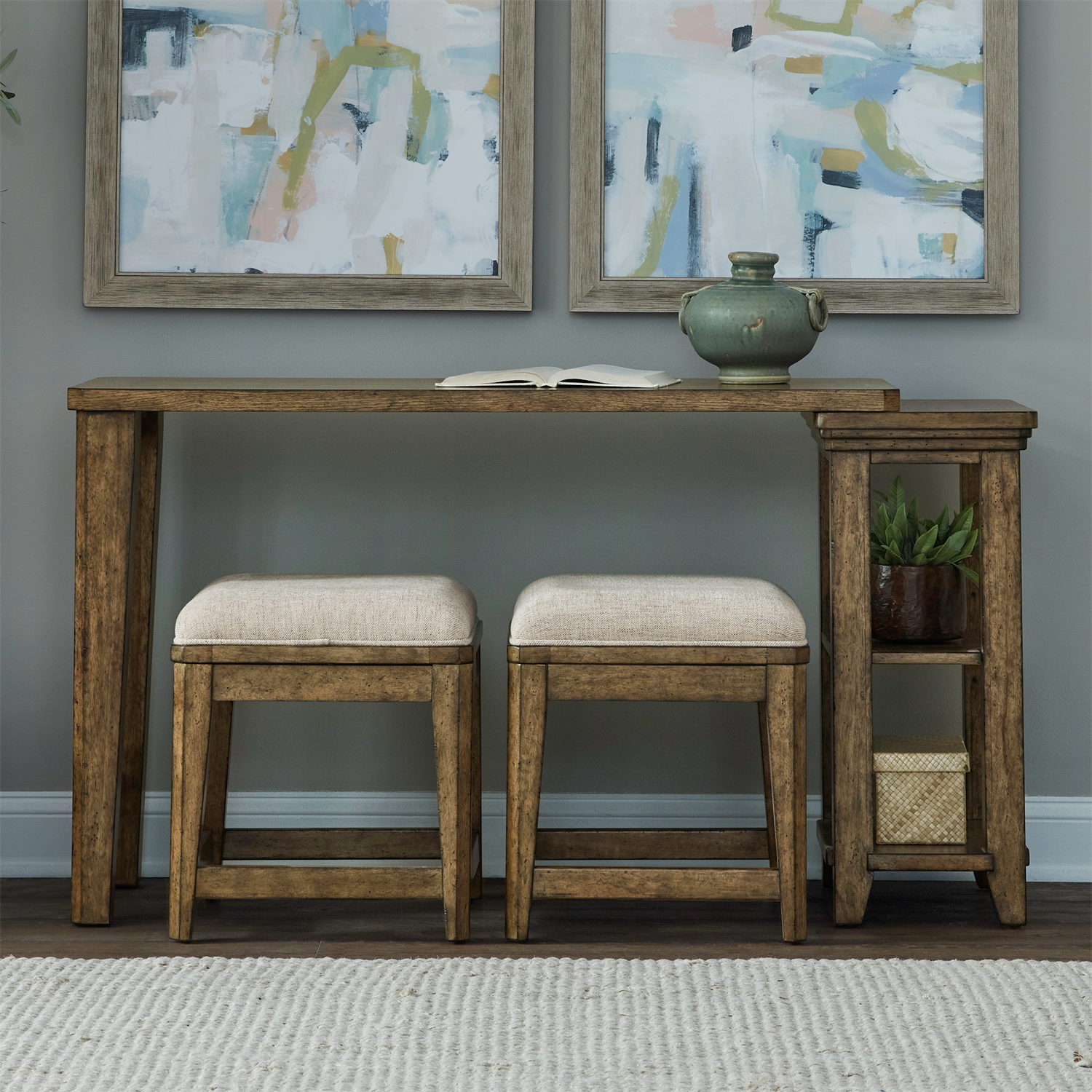 The Carolina Park Console Table and 2 Stools by Liberty Furniture offers elegant bookshelf storage, paired with two beige upholstered stools. Atop the wooden console table sits a green vase and an open book, while two abstract paintings above infuse artistic charm. A small side shelf holds a basket and another decorative pot, completing the look on a light rug for a cozy finish.