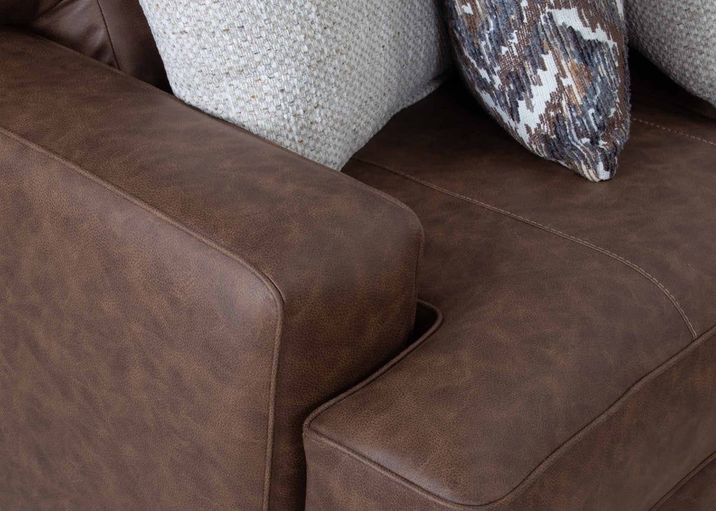 Close-up of the Asheville Duvall Vintage Brown Sectional by Franklin Furniture Corp., featuring two textured pillows—one in a solid light hue and the other with a vibrant geometric pattern. The sectional’s visible stitching and armrest details emphasize its quality craftsmanship and the comfort of its WOW Seating System.
