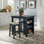 A kitchen featuring the Brook Creek Table and 2 Chairs 942NY-CD-3CTS by Liberty Furniture creates a cozy dining nook, complete with plush stools, open shelves displaying dishes, and a potted plant on the table. White cabinetry, stylish wall art, and a patterned rug enhance the bright atmosphere—making it an ideal solution for small space dining.