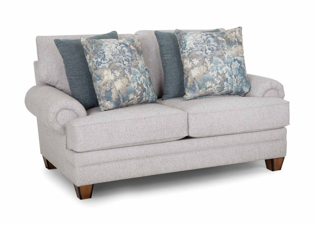 The Annisa Julien Cork Loveseat 95720-3574-20 by Franklin Furniture Corp. showcases a light gray upholstery with rolled arms, wooden legs, and the WOW Seating System. Its reversible seat and back cushions provide lasting comfort. This loveseat comes with four throw pillows—two in blue-gray and two featuring a blue floral pattern—and is displayed against a white background.