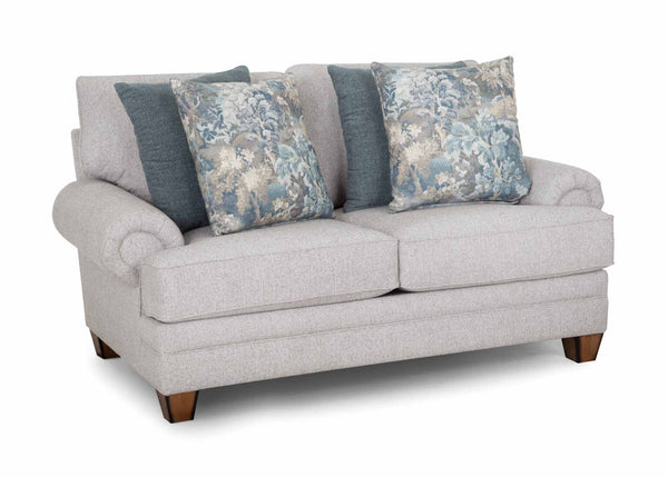 The Annisa Julien Cork Loveseat 95720-3574-20 by Franklin Furniture Corp. showcases a light gray upholstery with rolled arms, wooden legs, and the WOW Seating System. Its reversible seat and back cushions provide lasting comfort. This loveseat comes with four throw pillows—two in blue-gray and two featuring a blue floral pattern—and is displayed against a white background.