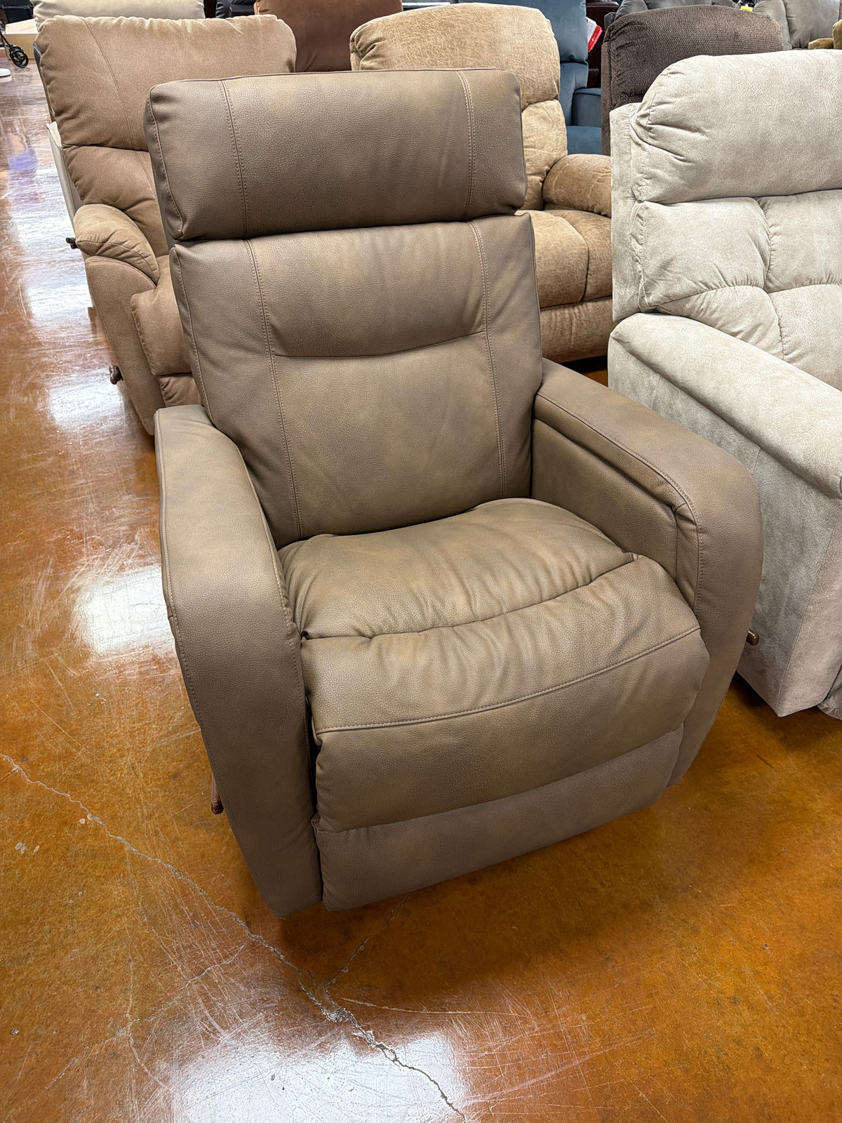 A taupe leather Baron Recliner 807-10-E2036-78 with an automotive-inspired design and a high backrest, positioned in a showroom with other recliners.