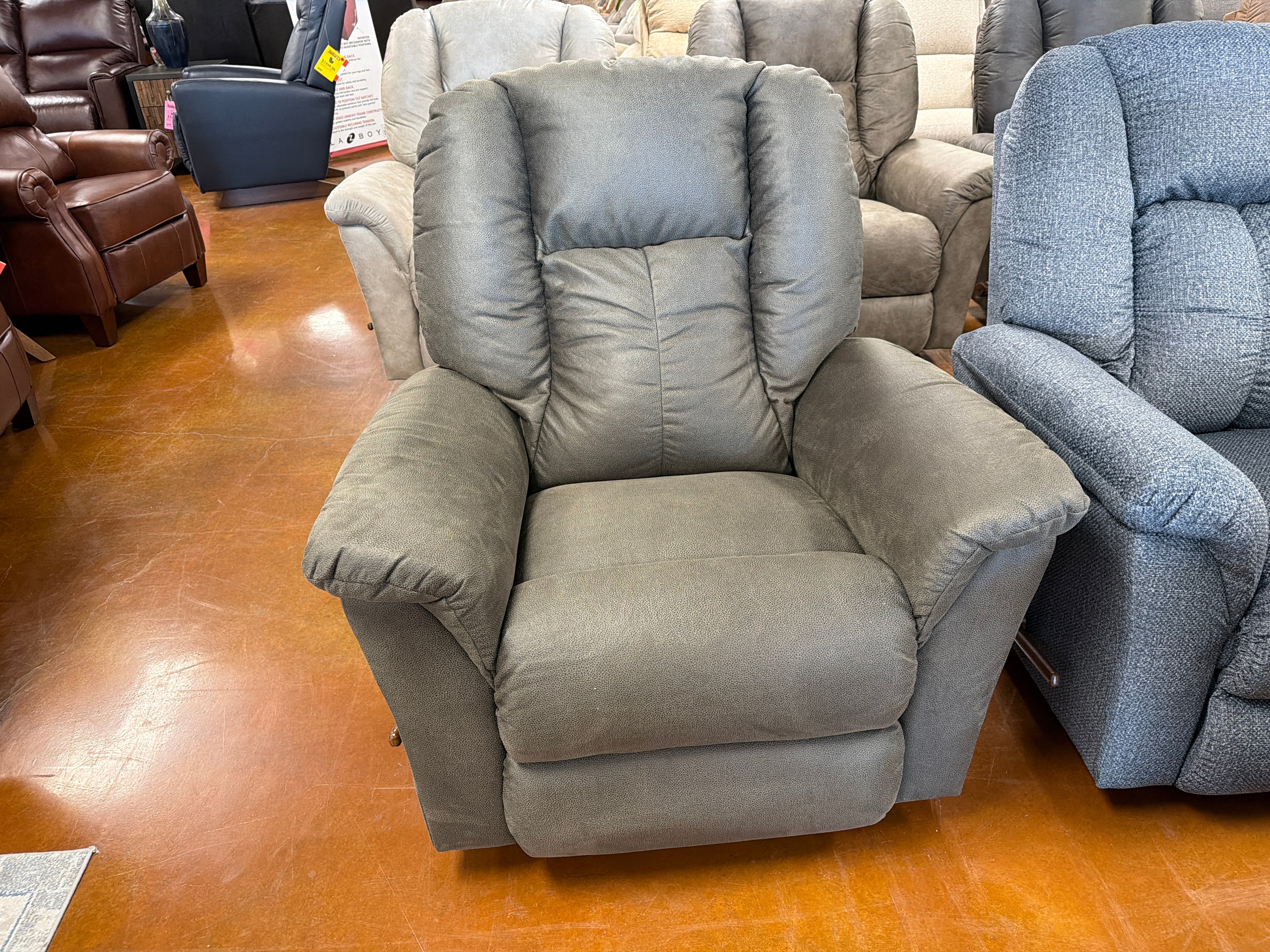 A gray upholstered La-Z-Boy Jasper Recliner 709-10-D1855-59 with plush cushions is displayed in a furniture store, surrounded by other recliners on a polished brown floor.