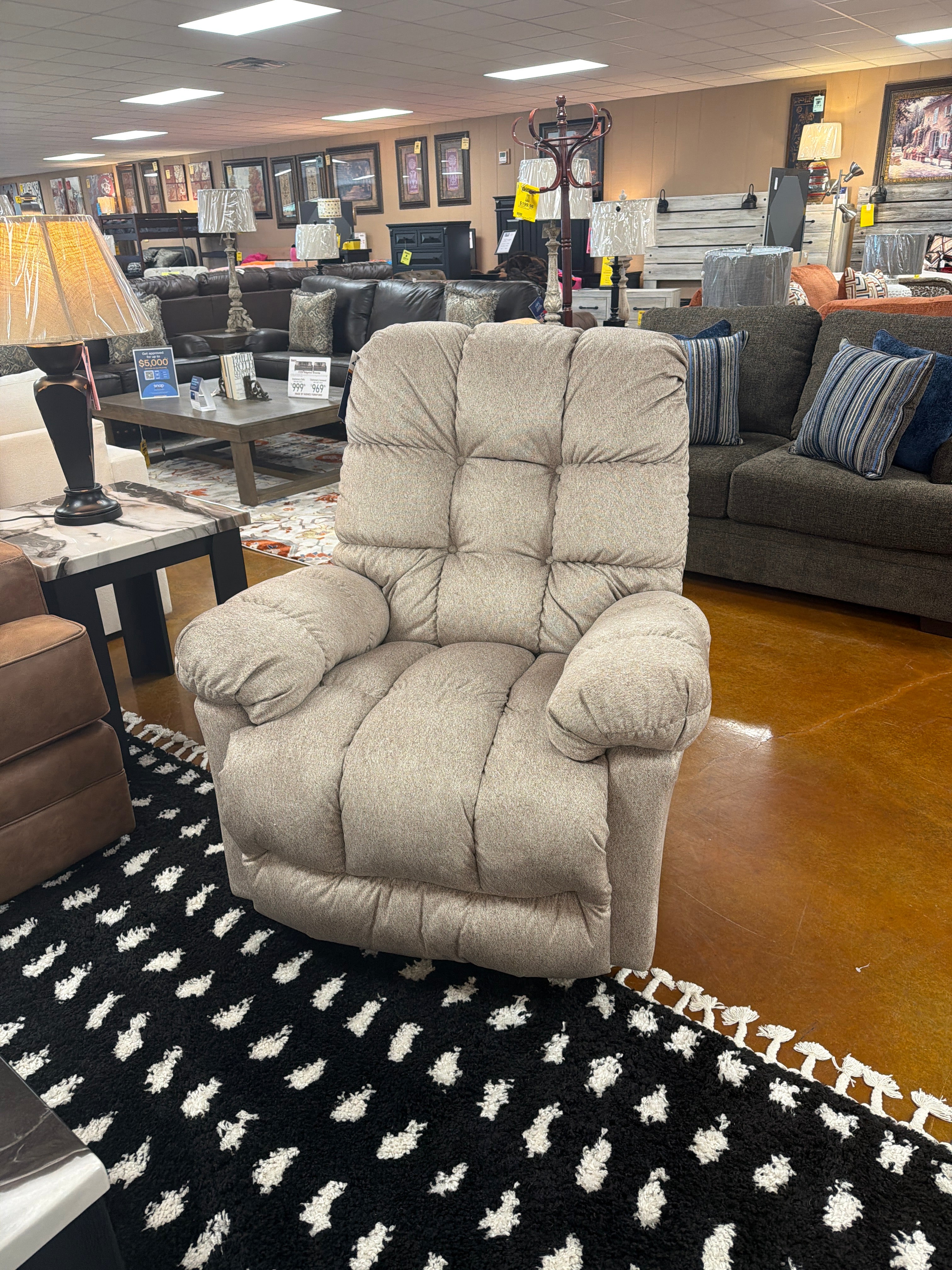 The Brosmer Beige Recliner 9MW87-1-20869 by Best Home Furnishings, featuring adjustable lumbar support and an overstuffed design, is displayed on a black and white patterned rug in a furniture store, surrounded by sofas, lamps, decor items, and illuminated by bright ceiling lights.