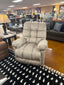 The Brosmer Beige Recliner 9MW87-1-20869 by Best Home Furnishings, featuring adjustable lumbar support and an overstuffed design, is displayed on a black and white patterned rug in a furniture store, surrounded by sofas, lamps, decor items, and illuminated by bright ceiling lights.