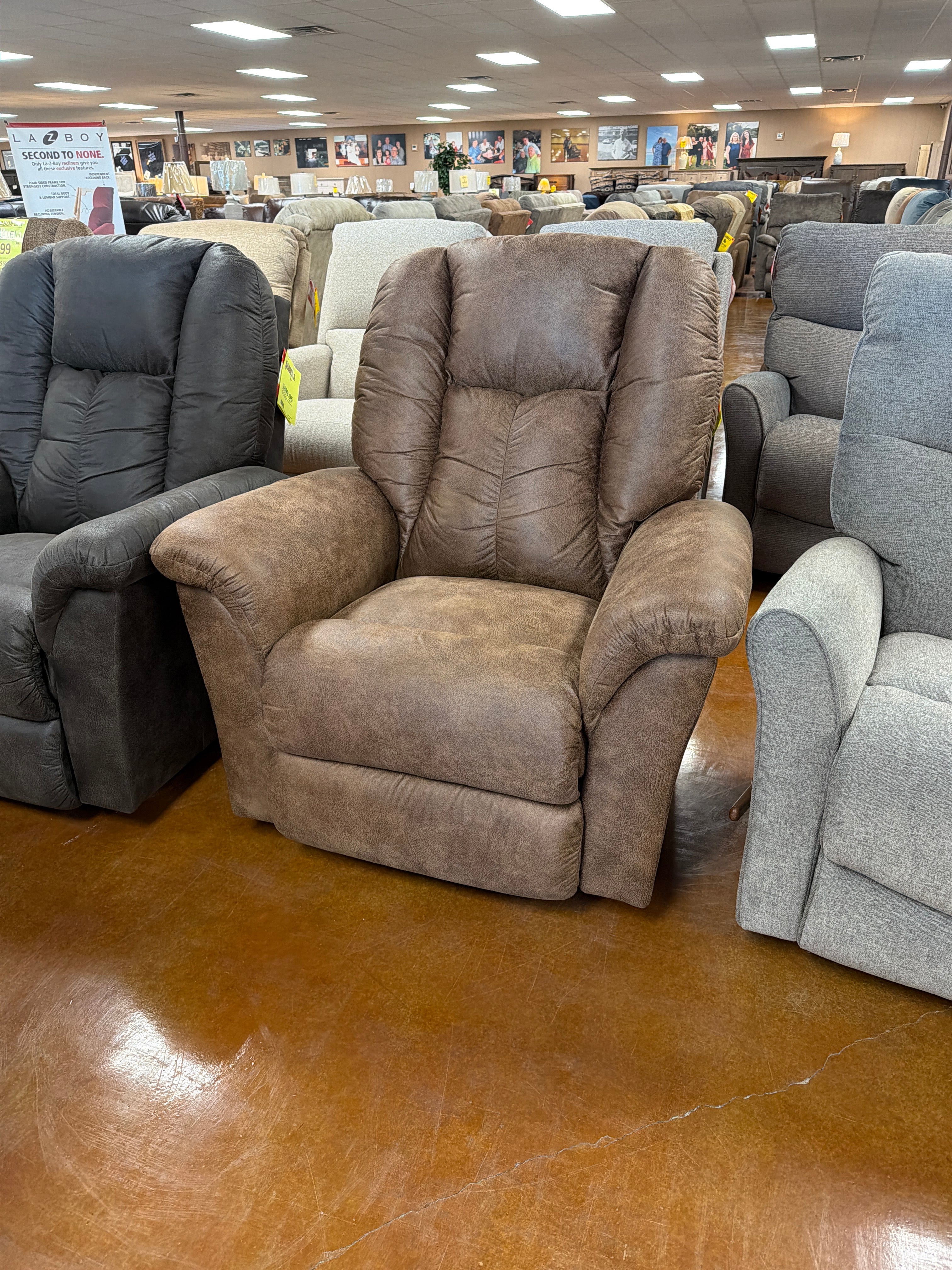 A La-Z-Boy Jasper Recliner (model 709-10-D1825-74) in brown upholstery is showcased in a furniture showroom, surrounded by other wide-seat recliners in different colors. The glossy brown floor and framed artwork on the walls provide a backdrop that complements the chair’s ultra-plush chaise seat.